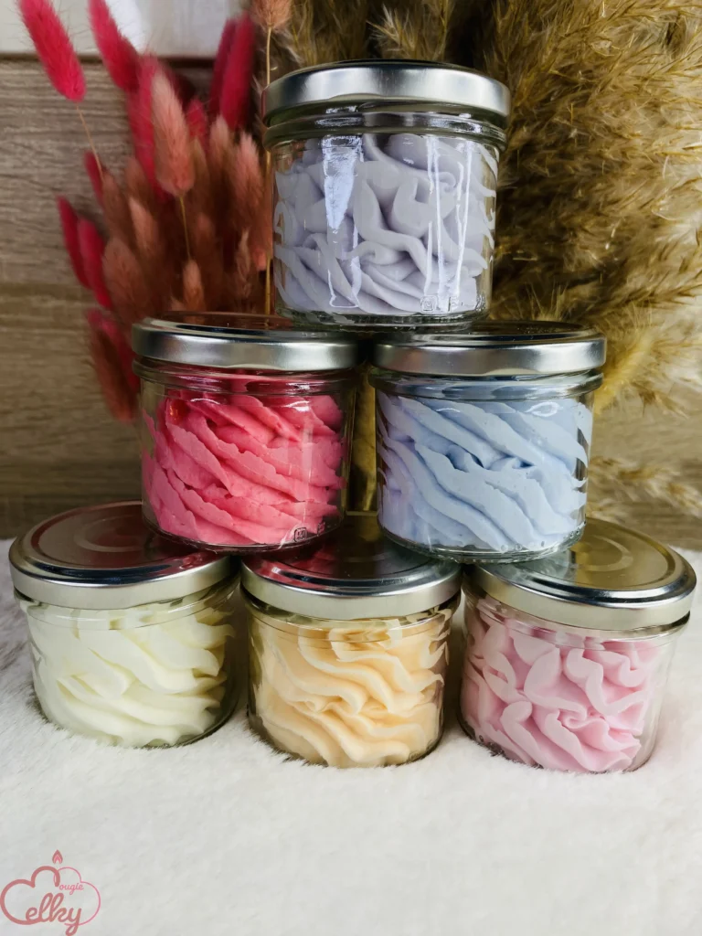 Six pots en verre contenant de la chantilly fondante parfumée aux couleurs pastel et vives, soigneusement disposés en pyramide. Chaque pot est garni d’une texture onctueuse rappelant une crème fouettée. En arrière-plan, un bouquet de fleurs séchées aux teintes naturelles et rosées ajoute une touche élégante et chaleureuse.
