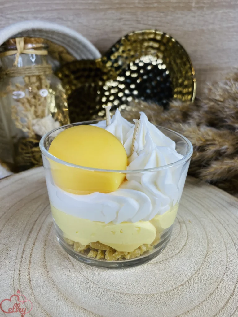 Bougie gourmande parfumée inspirée de la tarte citron meringuée, présentée dans une verrine avec une base biscuitée, une couche jaune rappelant la crème au citron et une décoration de chantilly en cire. Arrière-plan décoratif avec éléments dorés et fleurs séchées.