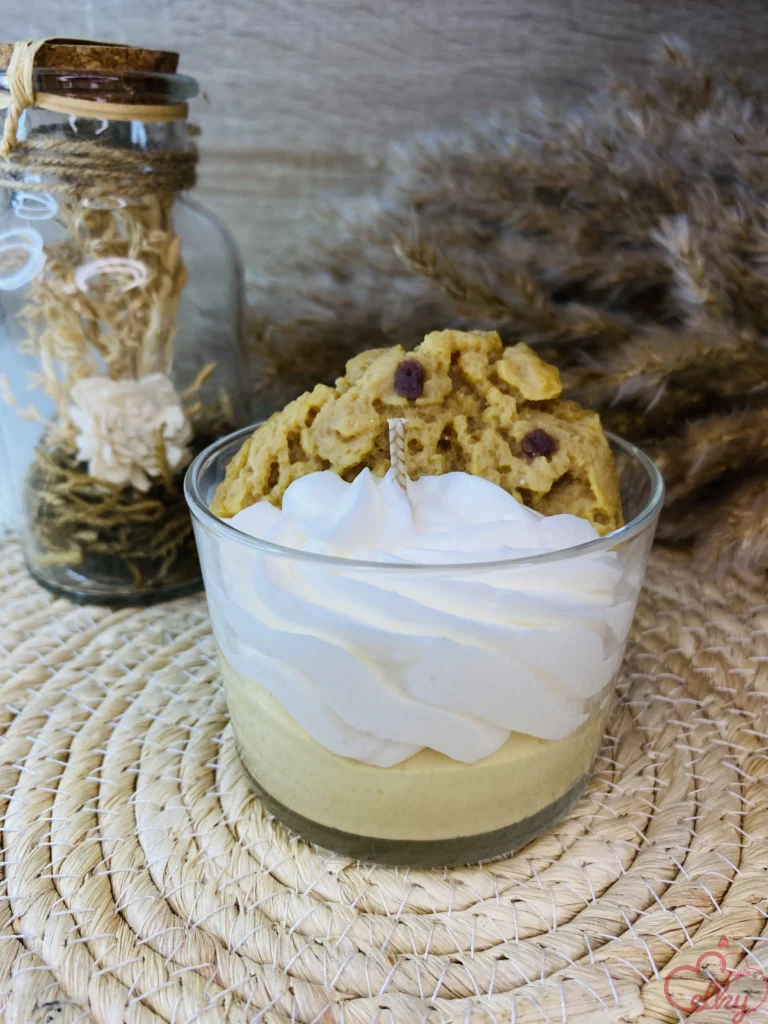 Bougie parfumée gourmande en forme de dessert, présentée dans un verre. Base crémeuse beige surmontée d’une mousse blanche façon chantilly et d’un biscuit façon cookie aux pépites de chocolat. En arrière-plan, une bouteille décorative avec des fleurs séchées et des éléments naturels.