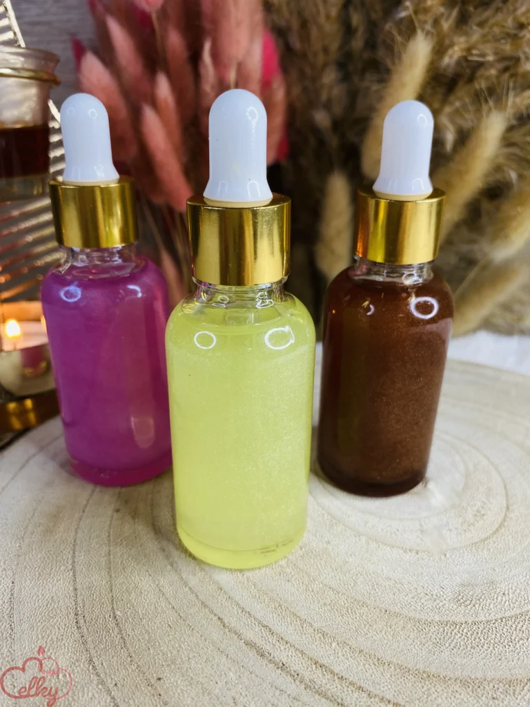 Flacons en verre contenant des huiles parfumées scintillantes aux couleurs vibrantes : rose, jaune et marron. Chaque flacon est équipé d’une pipette avec un embout blanc et un col doré, apportant une touche d’élégance. L’arrière-plan est décoré de fleurs séchées et d’une bougie allumée, créant une ambiance chaleureuse et raffinée.