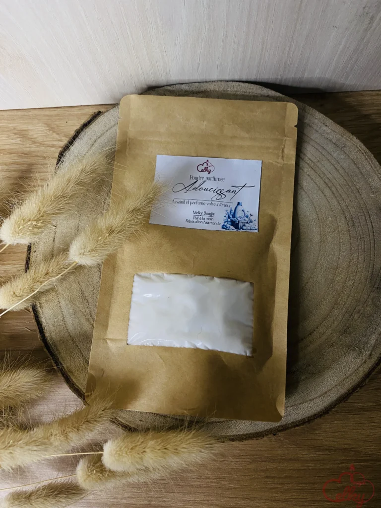 Sachet kraft de poudre parfumée pour intérieur, posé sur un support en bois avec des fleurs séchées en décoration. L'étiquette affiche l'inscription 'Adoucissant' et indique une fabrication en Normandie. Un sachet intérieur blanc est visible à travers une fenêtre transparente, suggérant un conditionnement soigné.