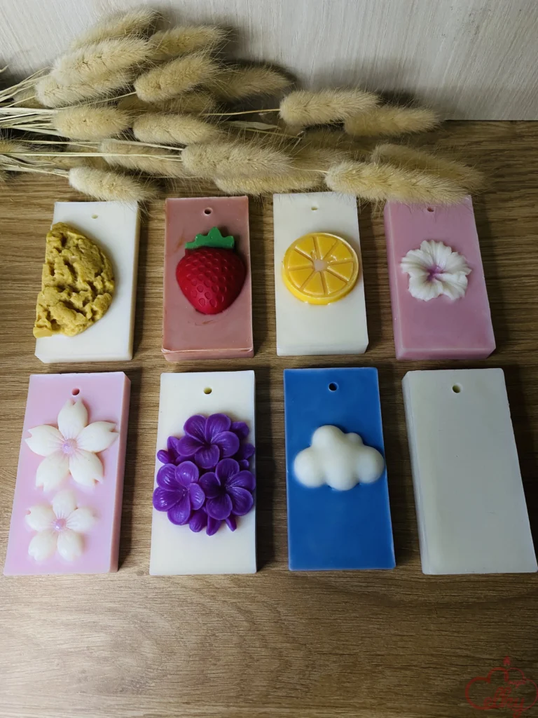Ensemble de huit suspensions parfumées rectangulaires en cire, ornées de divers motifs en relief (fleur, nuage, fruit, biscuit). Disposées sur une surface en bois, avec en arrière-plan un bouquet de lagurus séché, elles offrent une palette de couleurs variées : blanc, rose, bleu. Chaque suspension comporte un trou en haut pour y passer un lien d’accrochage.